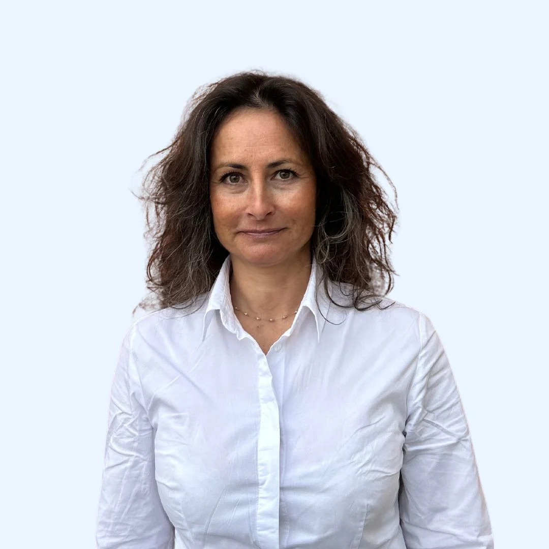 Une femme aux cheveux bruns ondulés, arborant un léger sourire, porte une chemise blanche boutonnée et se tient debout devant un fond clair et uni.