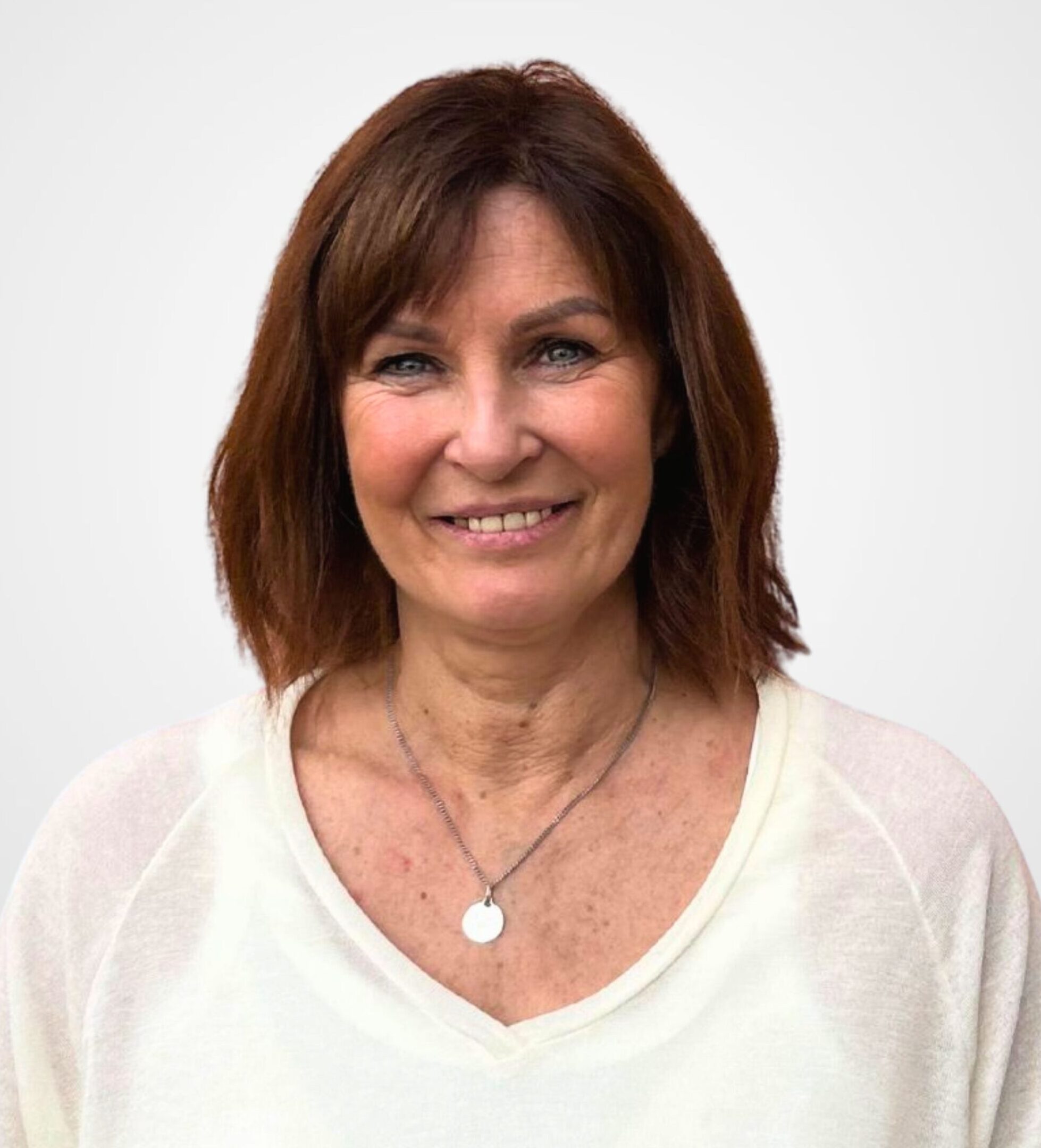 Une femme souriante aux cheveux châtain roux tombant jusqu'aux épaules, vêtue d'une chemise blanche et portant un collier avec un pendentif rond en argent.