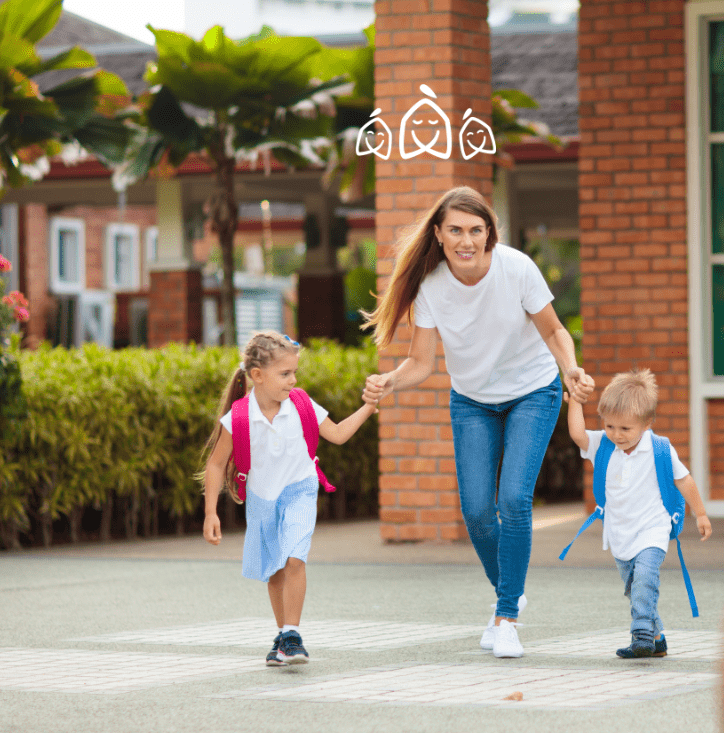 Une femme marche main dans la main avec une jeune fille et un jeune garçon, tous portant des vêtements décontractés et des sacs à dos, à l'extérieur d'un bâtiment scolaire avec des colonnes en briques et de la verdure.