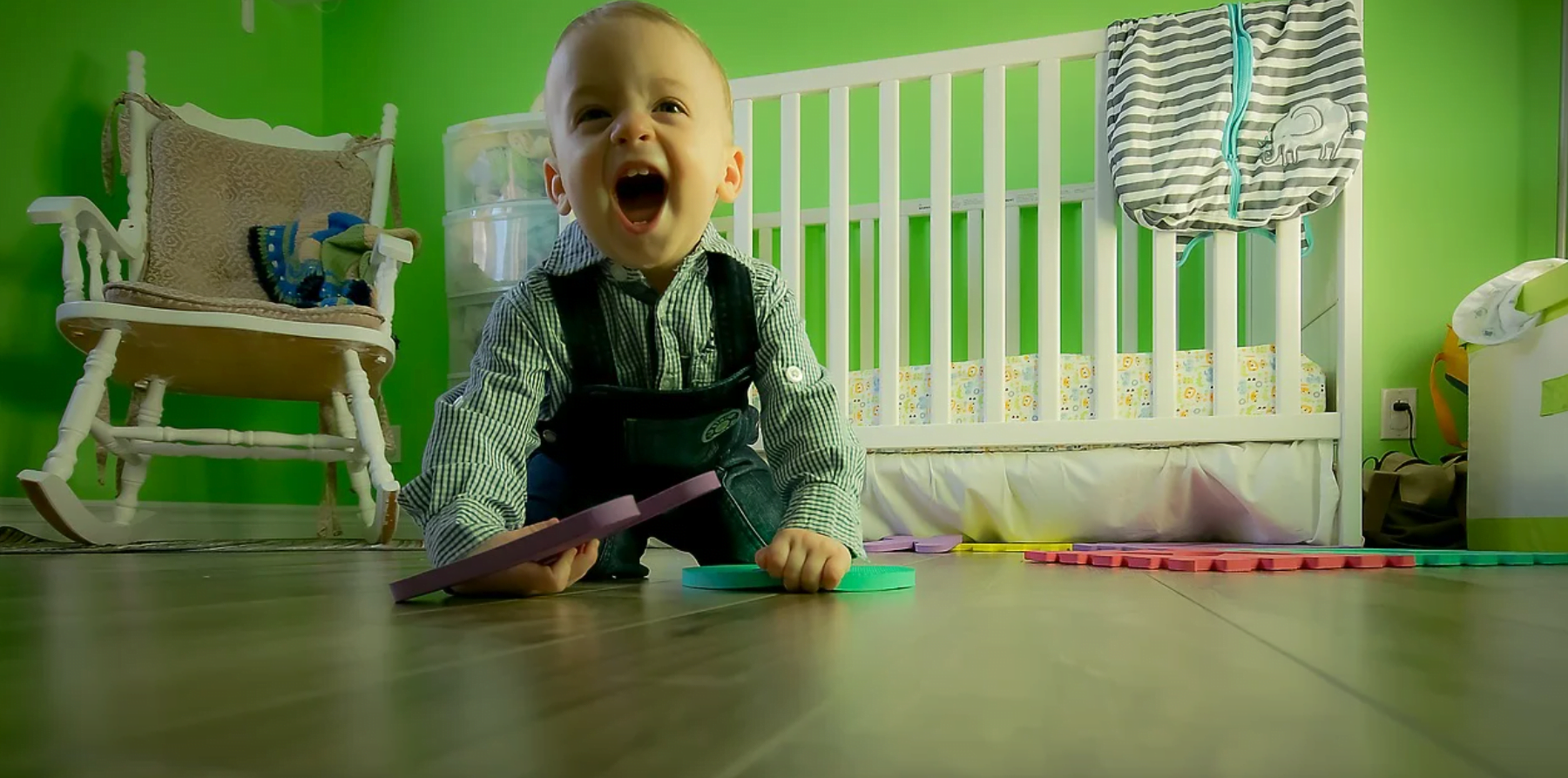 Un bambin souriant portant des bretelles rampe sur un plancher en bois, tenant des pièces de puzzle en mousse colorées, dans une chambre d'enfant lumineuse avec un berceau blanc et des murs verts, surveillée de près par une aide à domicile attentive.