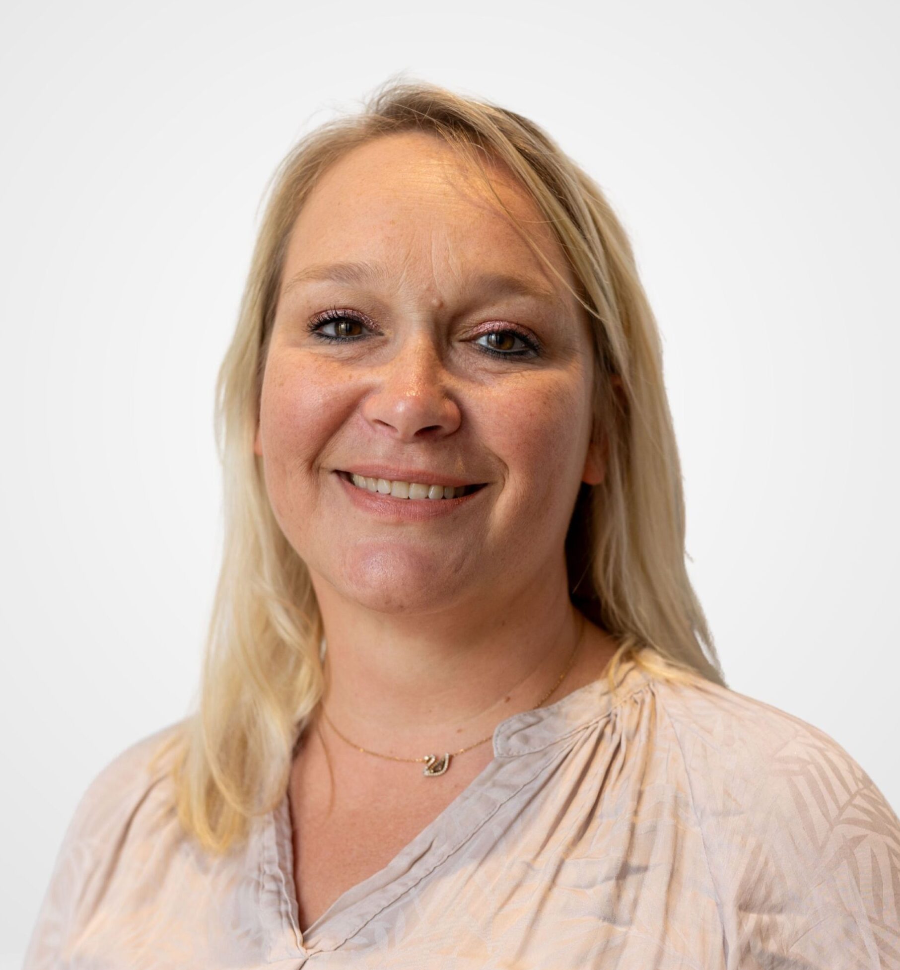 Une femme souriante aux cheveux blonds, vêtue d'un chemisier de couleur claire et portant un collier raffiné.