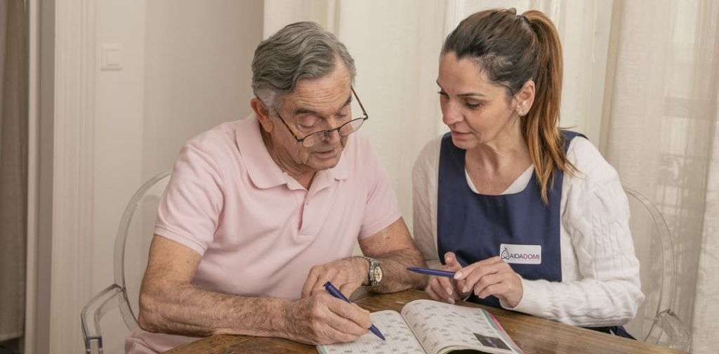 Un homme âgé et une aide-soignante d'aidadomi sont assis à une table et regardent ensemble un livre de puzzle. L'homme tient un stylo tandis que l'aide-soignante, qui porte un badge, lui montre gentiment le livre pour l'aider.