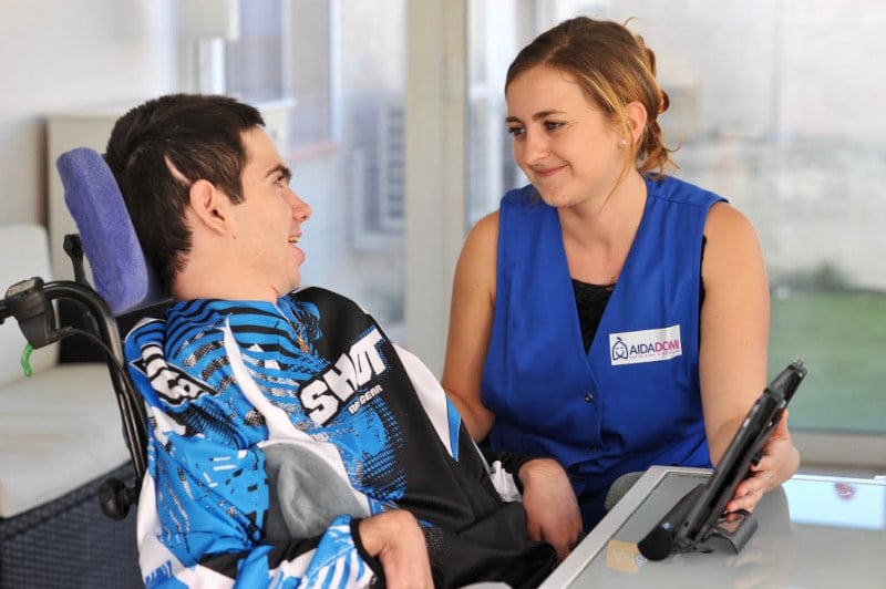 Un jeune homme en fauteuil roulant sourit à une femme en gilet bleu qui tient une tablette. À l'intérieur, elle fournit une aide éducative aux adolescents handicapés, en l'assistant ou en conversant avec lui.