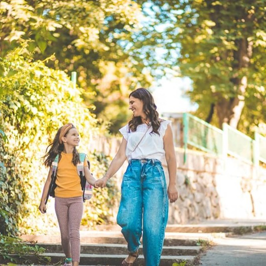 Une femme et une jeune fille se tiennent par la main, se sourient et se promènent à l'extérieur par une journée ensoleillée. La jeune fille porte un sac à dos, ce qui laisse supposer qu'elles se rendent à l'école ou en reviennent. Des arbres et de la verdure se trouvent à l'arrière-plan.