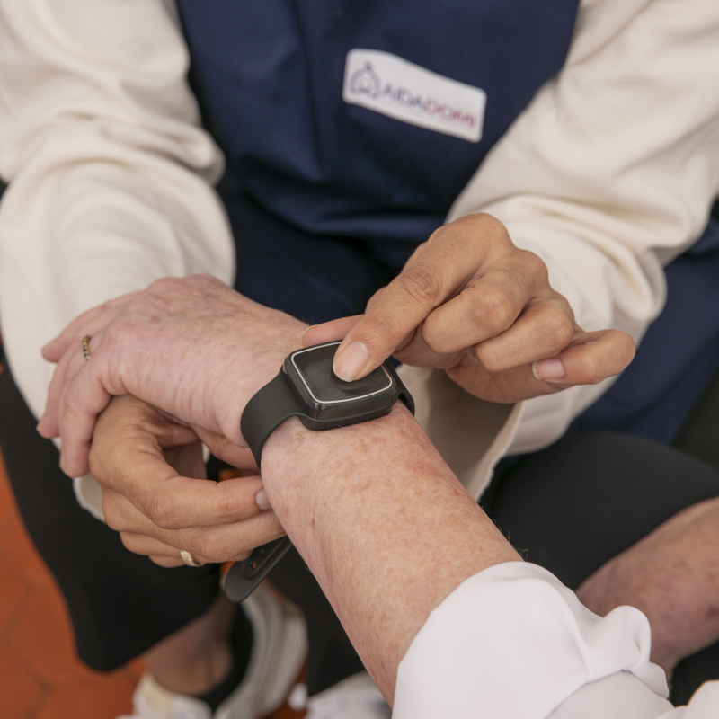 Un aidant aide une personne âgée à utiliser une smartwatch pour la téléassistance à domicile, en touchant doucement l'écran pendant que la montre est portée au poignet de la personne âgée. L'aidant porte un uniforme avec un badge visible, mettant l'accent sur la sécurité.
