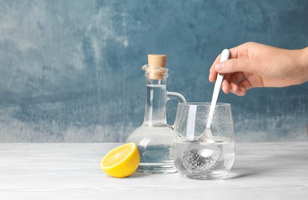 Une main remue une cuillère dans un verre de liquide transparent, avec une carafe de vinaigre blanc salle de bain et un citron coupé en deux sur une surface blanche sur un fond bleu texturé.