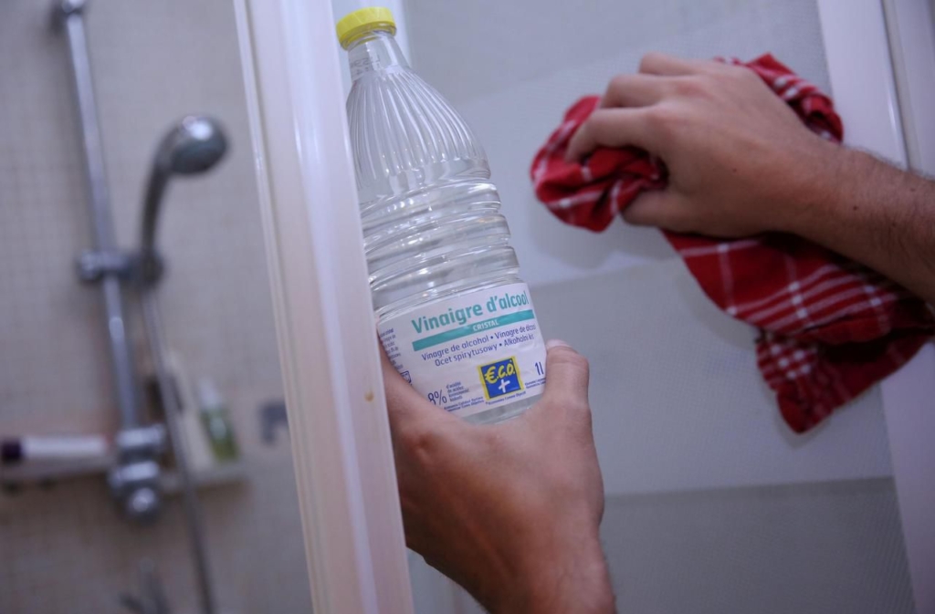 Une personne nettoie une porte de douche avec un chiffon rouge dans une main et une bouteille de vinaigre blanc salle de bain dans l'autre, soulignant son efficacité pour un entretien impeccable de la salle de bain.