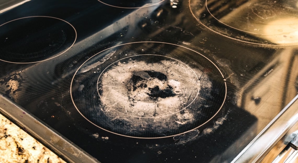 Une cuisinière en verre présentant des taches de brûlé, de graisse et de résidus visibles, en particulier sur le brûleur central. La surface semble sale et a besoin d'un nettoyage naturel plaque cuisson. Une partie d'un comptoir en granit est visible sur le côté gauche.