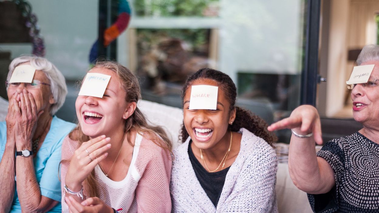 Quatre personnes d'âges différents sont assises ensemble, riant, chacune avec une note autocollante sur le front alors qu'elles s'amusent à jouer à des jeux maison sans accessoires. Une femme fait des gestes animés, ce qui ajoute à la vivacité et à la joie de la scène.