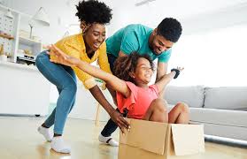 Une enfant souriante est assise dans une boîte en carton, les bras tendus, tandis que deux adultes, également souriants, la poussent et la guident dans une salle de séjour lumineuse et joyeuse - un parfait exemple de jeux sans matériel.