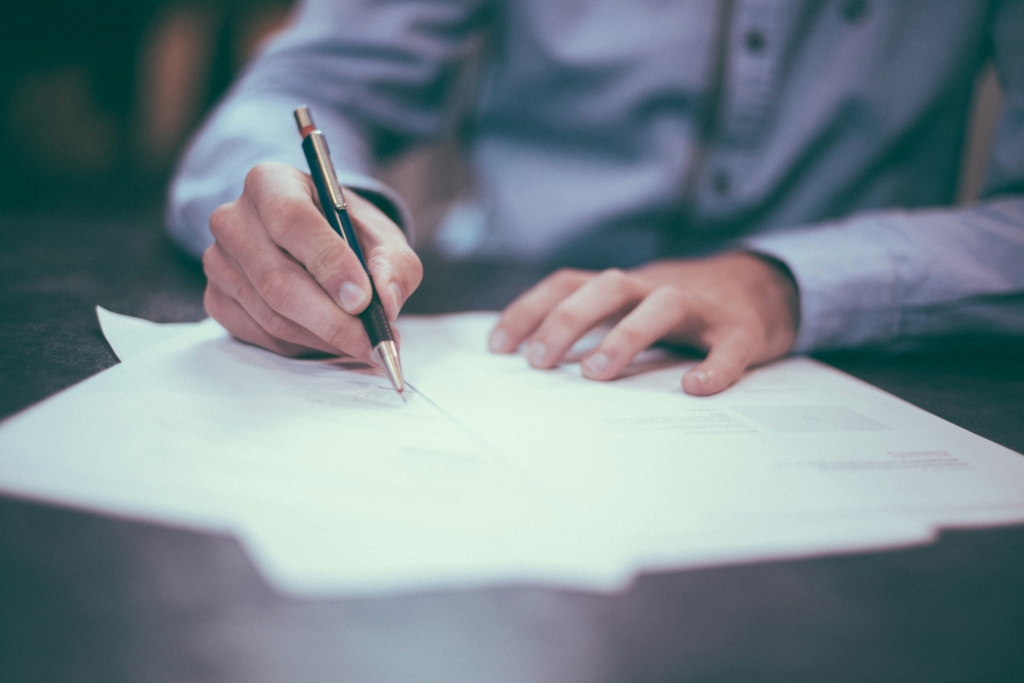 Une personne portant une chemise bleue écrit au stylo sur des feuilles de papier au sujet des services à la personne -50%, l'accent étant mis sur ses mains et le stylo, tandis que l'arrière-plan reste flou.