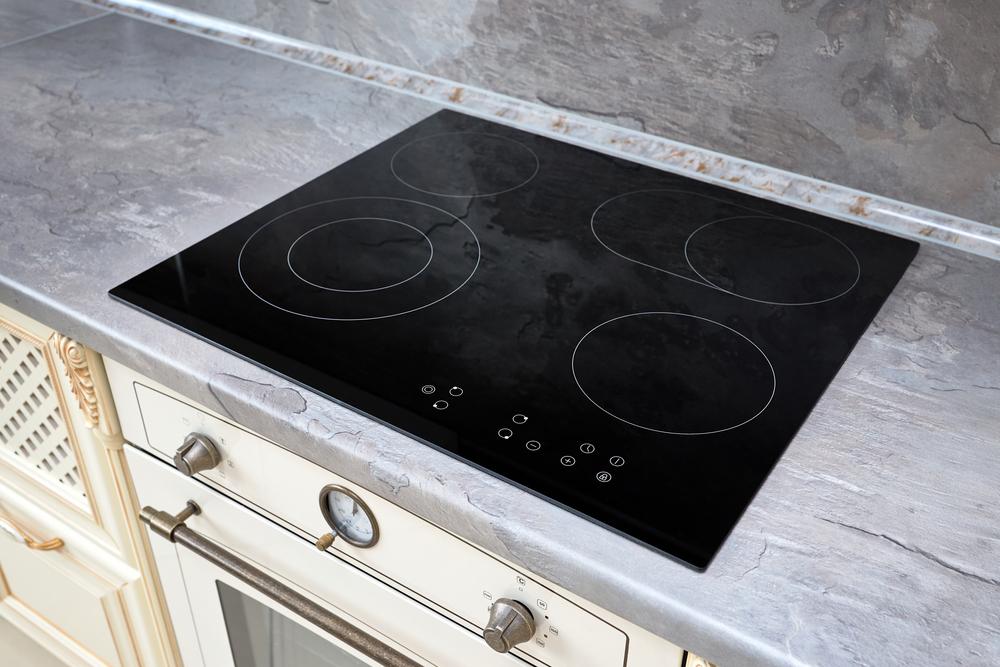 Une cuisinière électrique moderne en verre noir avec quatre brûleurs circulaires est encastrée dans un comptoir en marbre gris au-dessus d'un four blanc, doté d'une poignée argentée et d'une horloge analogique ronde - parfait pour ceux qui préfèrent le nettoyage naturel plaque cuisson.