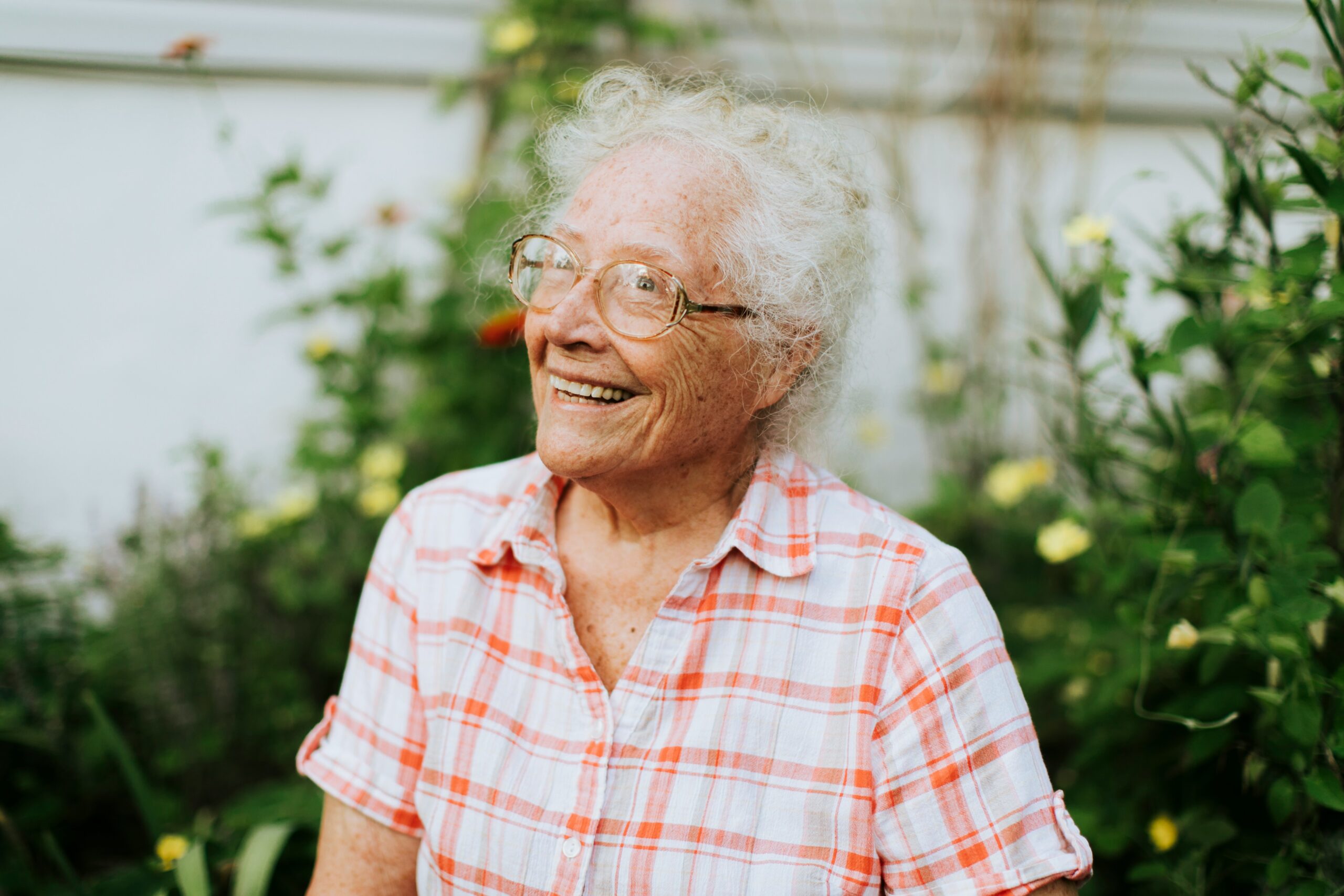 Une femme âgée aux cheveux blancs bouclés et aux lunettes sourit, vêtue d'une chemise à carreaux rouges et blancs, debout dans un jardin avec des plantes vertes et des fleurs jaunes en arrière-plan.