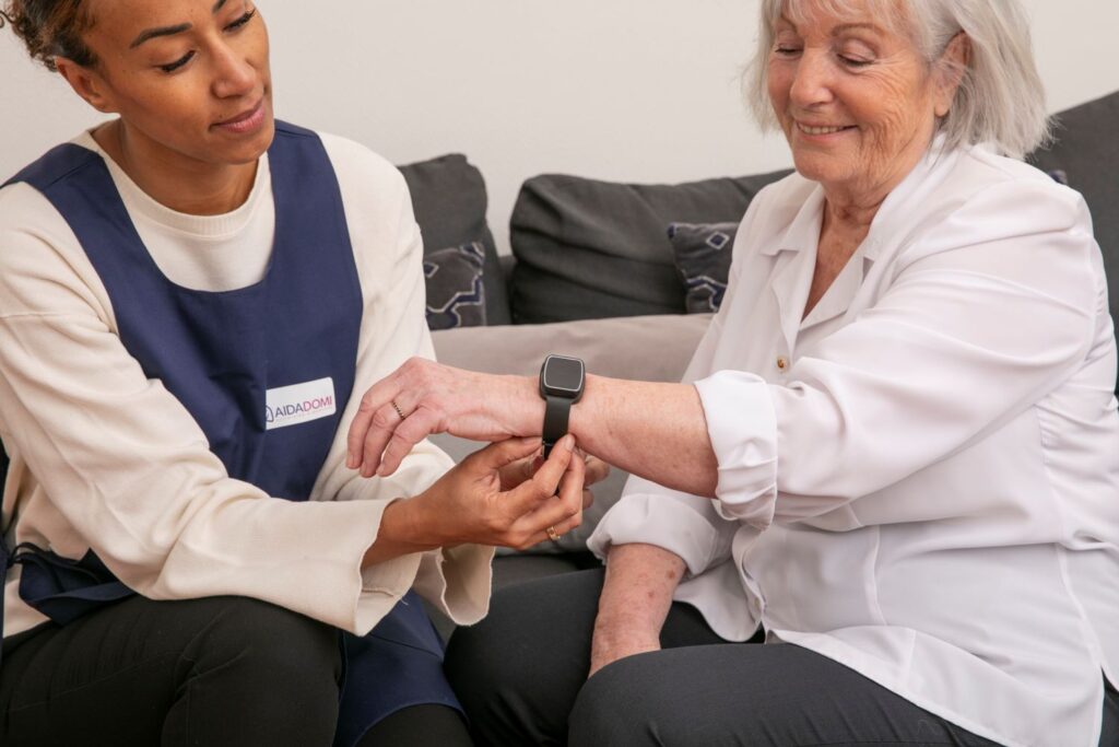 Une aide-soignante aide une femme âgée à mettre une montre intelligente pour la téléassistance à domicile, alors qu'elles sont assises ensemble sur un canapé. La femme âgée sourit, et tous deux semblent à l'aise et engagés.