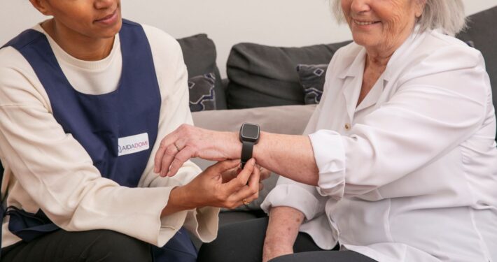 Une aide-soignante aide une femme âgée à mettre une montre intelligente pour la téléassistance à domicile, alors qu'elles sont assises ensemble sur un canapé. La femme âgée sourit, et tous deux semblent à l'aise et engagés.