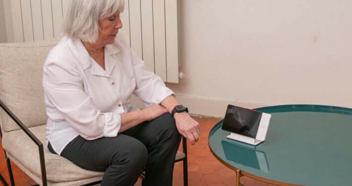 Une femme âgée aux cheveux grisonnants est assise sur une chaise et regarde un appareil numérique posé sur une table basse en verre vert devant elle. Cette scène reflète la téléassistance à domicile moderne pour une sécurité et une tranquillité d'esprit accrues.