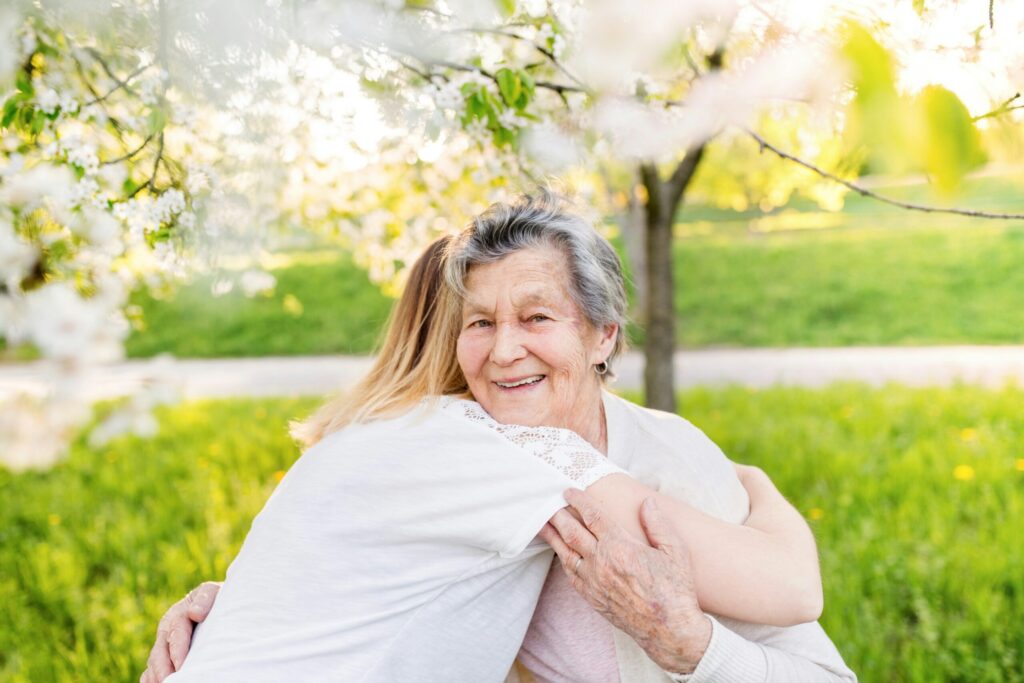 Une femme âgée sourit en étreignant une personne plus jeune dans un parc verdoyant et ensoleillé, entouré d'arbres en fleurs blanches.