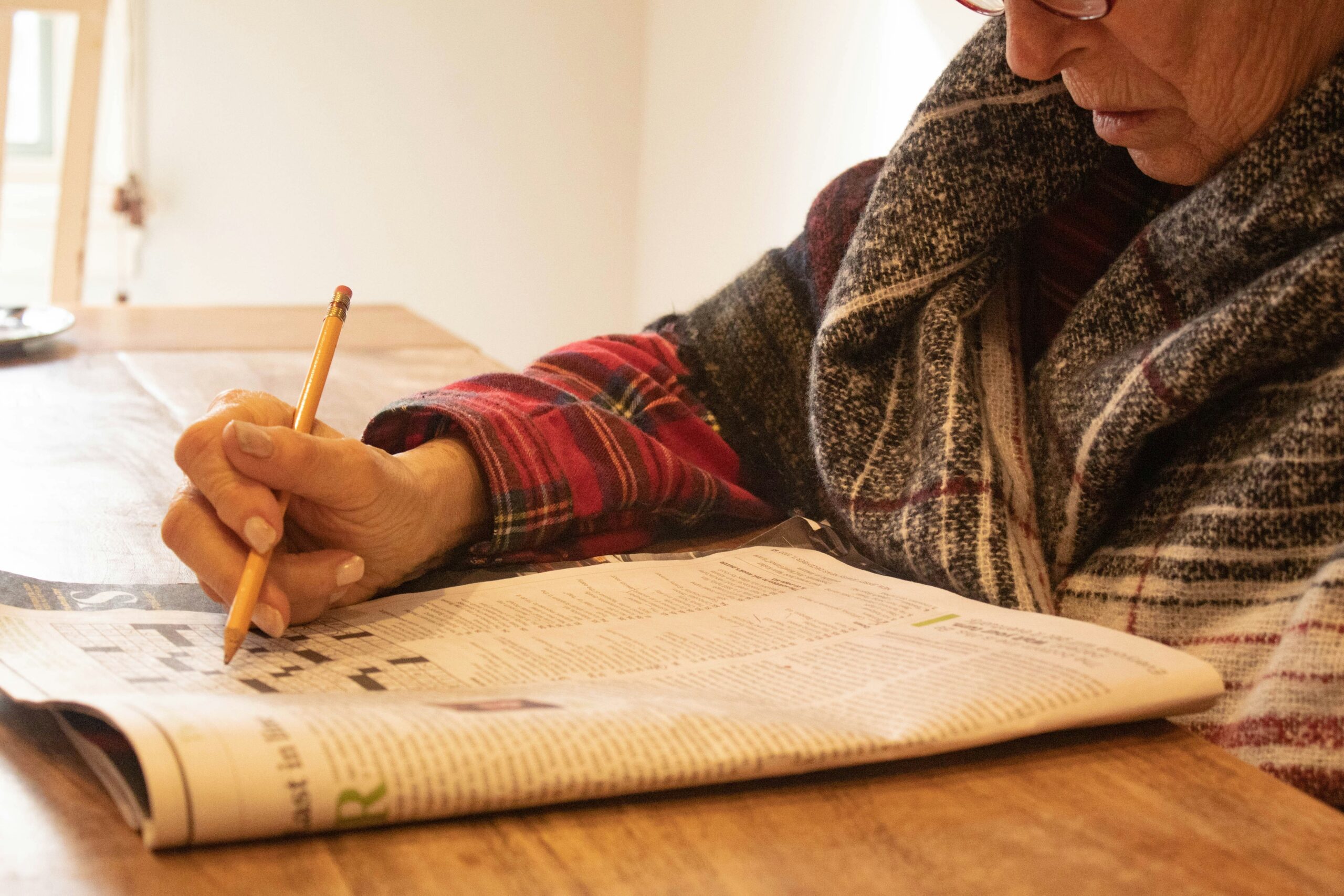 Une personne portant des lunettes et une chemise à carreaux est assise à une table en bois, concentrée à remplir une grille de mots croisés dans un journal, peut-être en train de faire une pause après avoir été aide ménagère du CESU.