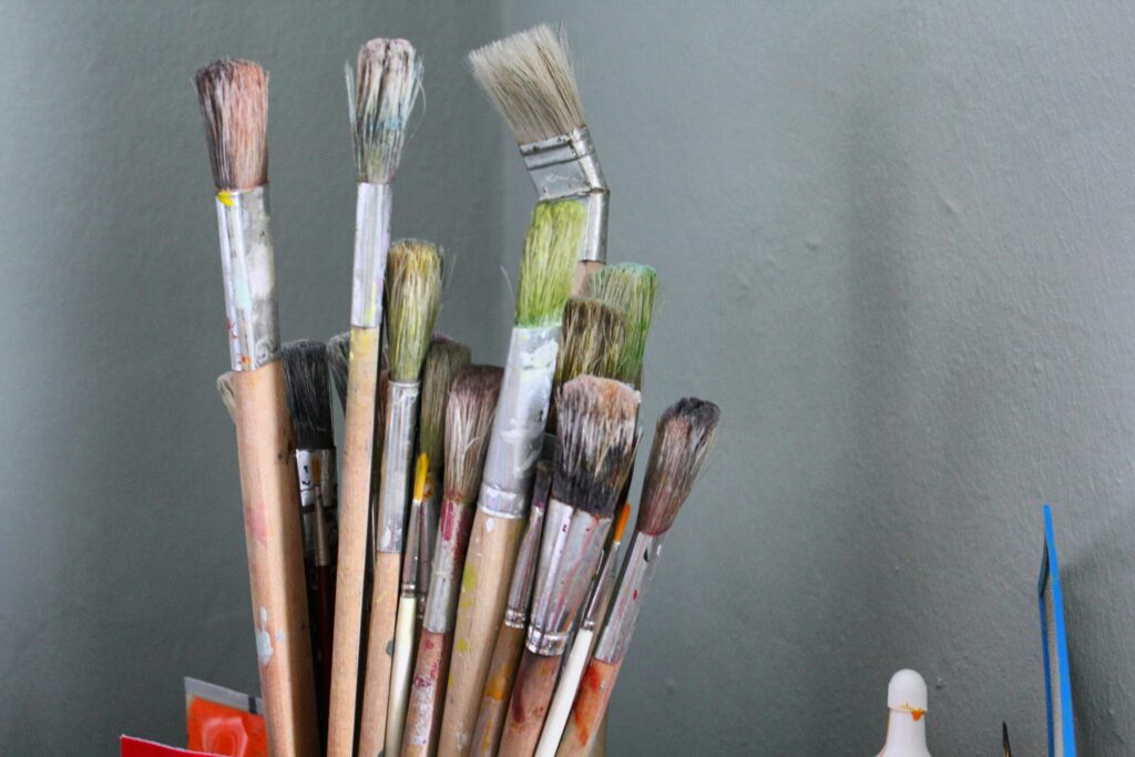 Une collection de pinceaux usagés aux manches tachés et aux poils colorés se tient debout dans un conteneur contre un mur gris uni.