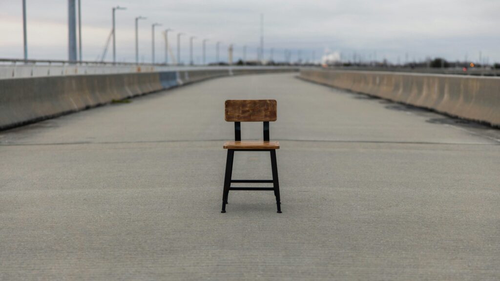 Une simple chaise en bois avec un dossier se trouve au milieu d'une large route en béton vide, entourée de barrières et de lampadaires par une journée nuageuse.