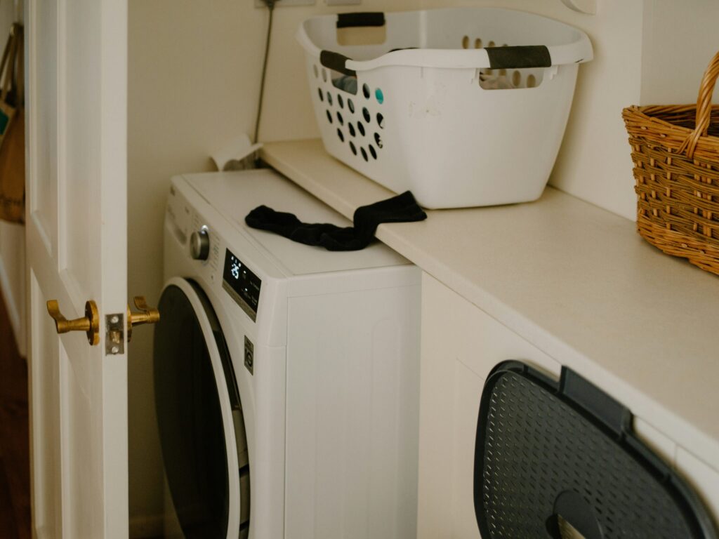 Une buanderie avec une machine à laver à chargement frontal, un panier à linge blanc sur le comptoir, un panier en osier et une chaussette noire drapée sur le bord du comptoir. La porte de la pièce est partiellement ouverte.