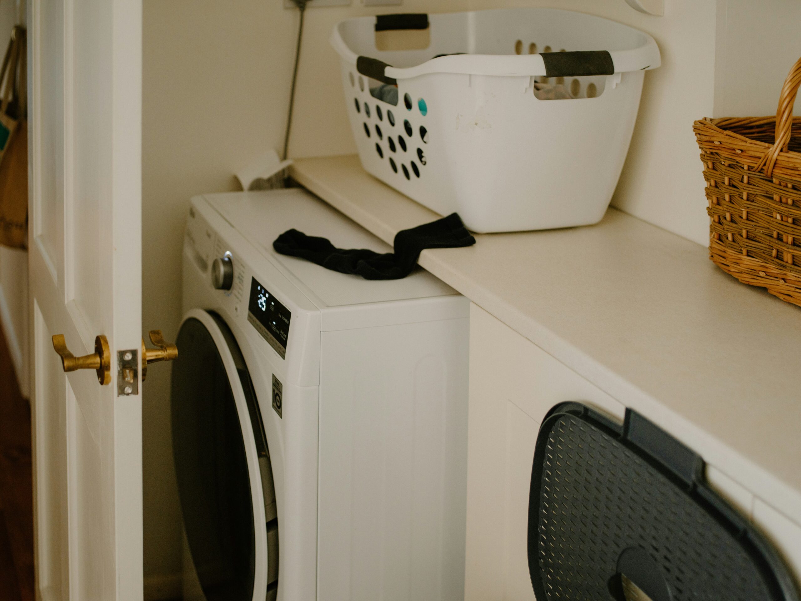 Une buanderie avec une machine à laver à chargement frontal, un panier à linge blanc sur le comptoir, un panier en osier et une chaussette noire drapée sur le bord du comptoir. La porte de la pièce est partiellement ouverte.