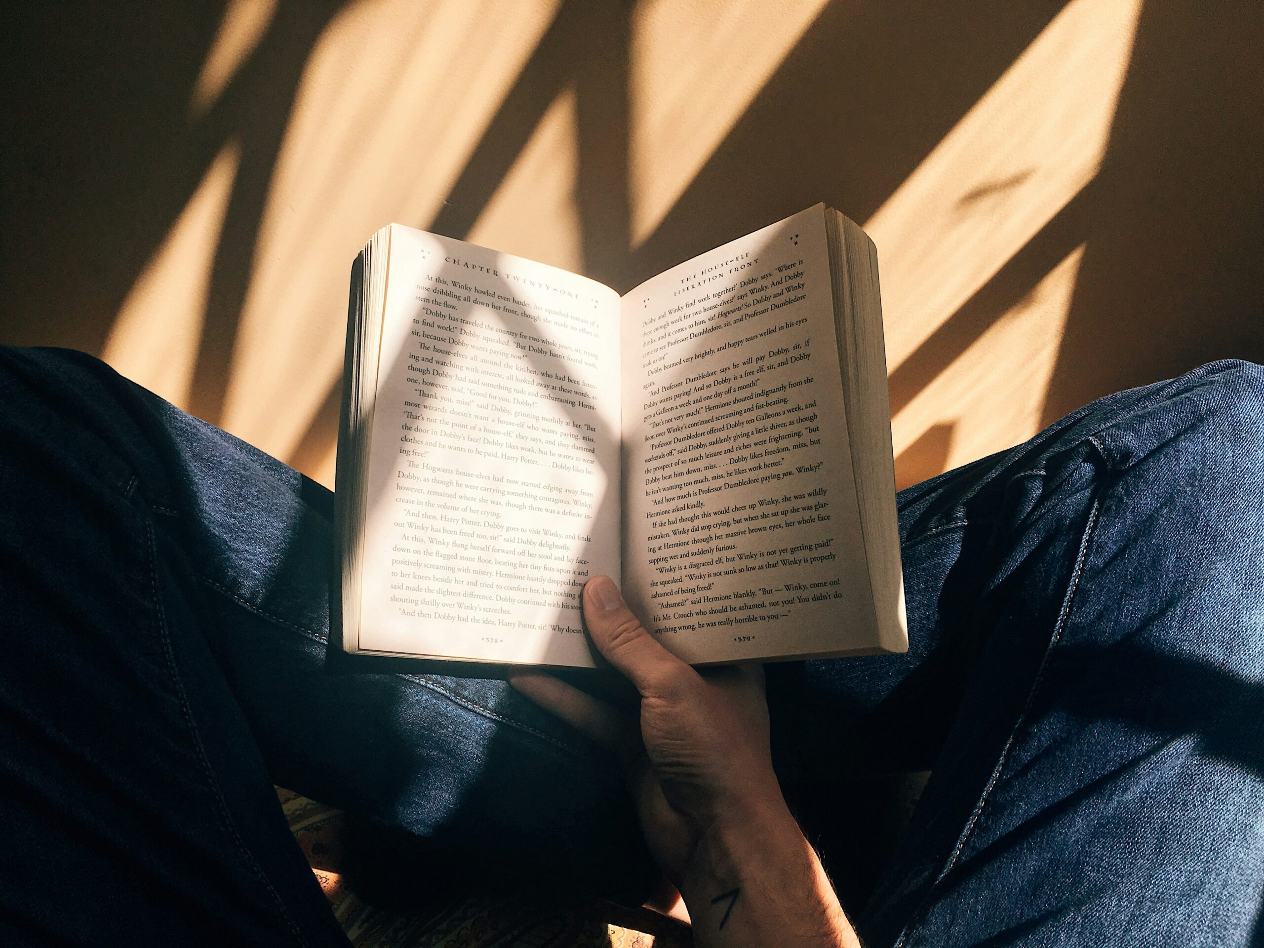 Une personne portant un jean bleu est assise les jambes croisées, tenant un livre ouvert sur ses genoux. La lumière du soleil et les ombres tombent sur les pages du livre et sur ses jambes.
