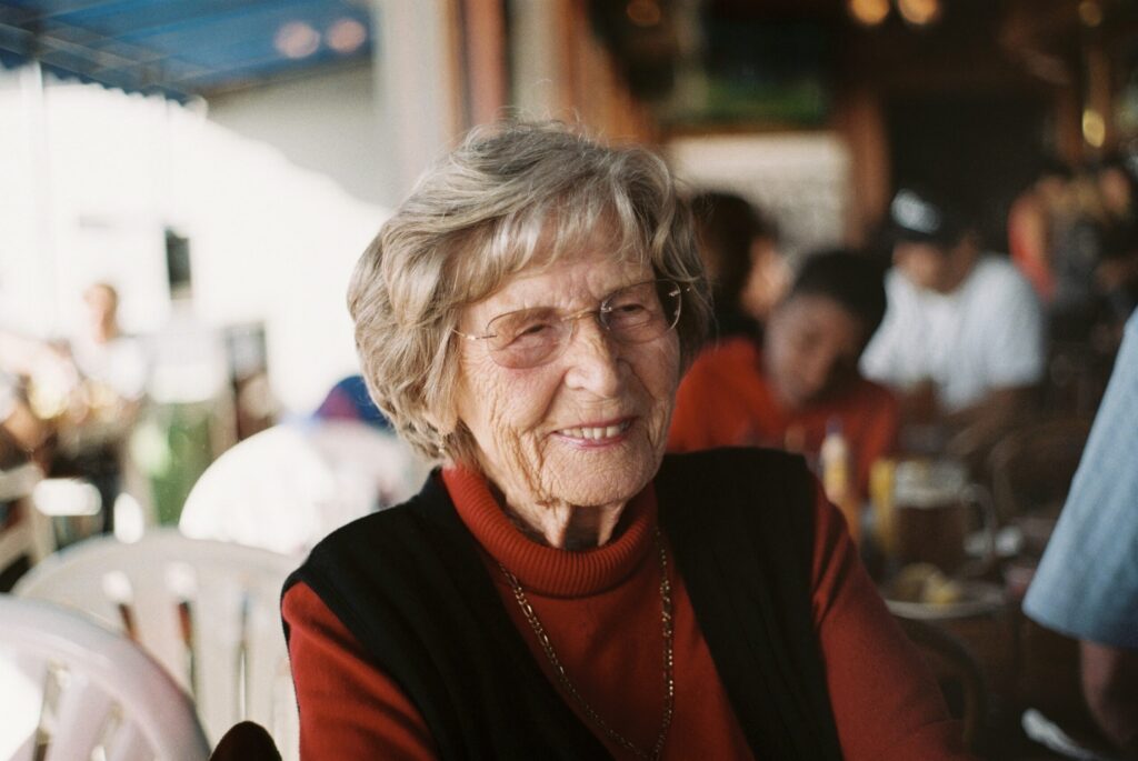 Une femme âgée aux cheveux gris courts et aux lunettes sourit alors qu'elle est assise dans un café intérieur très fréquenté. Son col roulé rouge et son gilet noir sont impeccables, peut-être grâce à l'aide ménagère de la CESU qui l'aide dans ses tâches ménagères. D'autres clients discutent en arrière-plan.