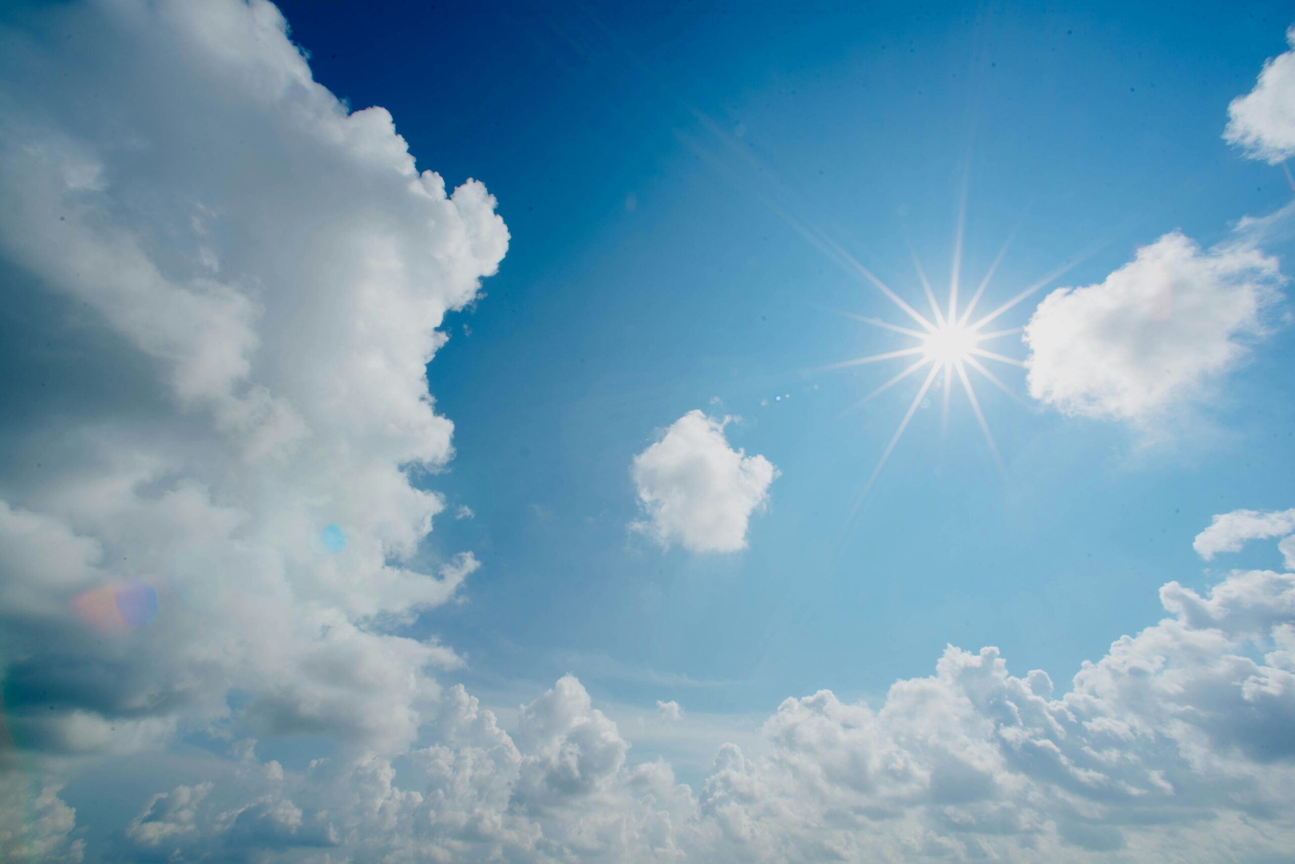 Soleil brillant dans un ciel bleu clair avec des nuages blancs épars, certains paraissant cotonneux et larges, créant une scène diurne vibrante et sereine.