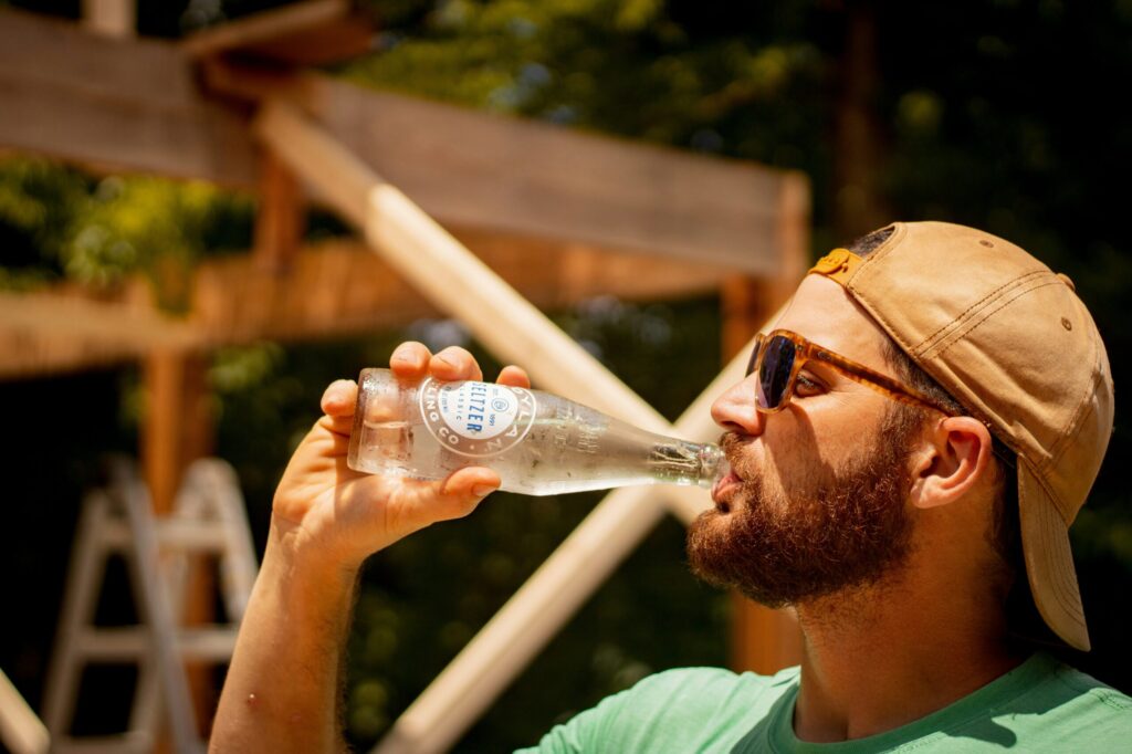 Un homme portant des lunettes de soleil et une casquette à l'envers boit une bouteille en verre transparent en plein air, avec des poutres en bois et une échelle en arrière-plan, par une journée ensoleillée.