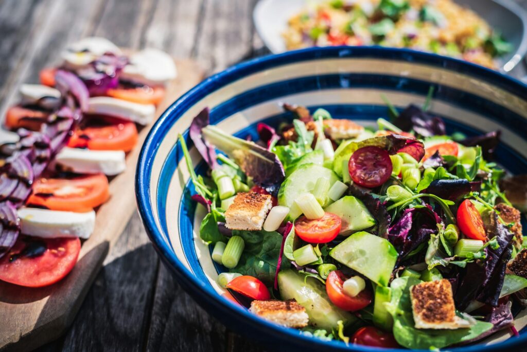 Une salade colorée avec des tomates cerises, des concombres, des légumes verts, des croûtons et des oignons verts dans un bol à rayures bleues, avec des tranches de tomates, de mozzarella et d'oignons sur une planche en bois à l'arrière-plan.