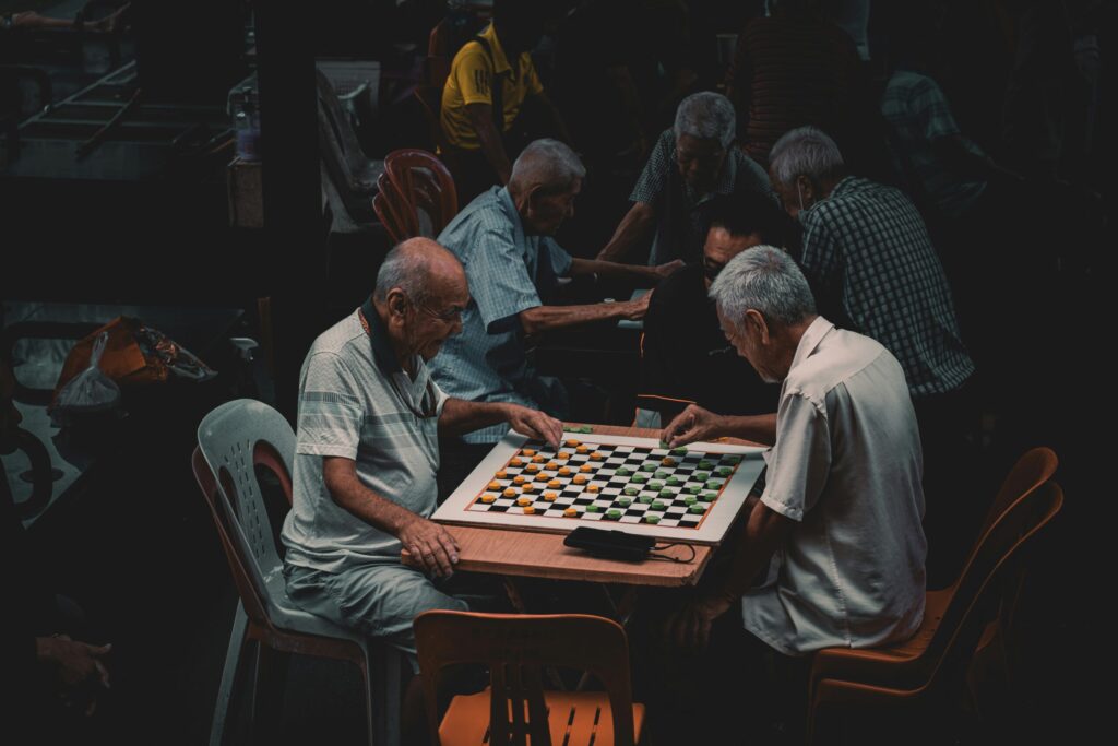 Deux hommes âgés sont assis l'un en face de l'autre à une petite table, jouant intensément à un jeu avec un plateau à carreaux, tandis que d'autres hommes - certains assistés par une aide ménagère du CESU - se rassemblent et jouent à des jeux dans l'arrière-plan faiblement éclairé.