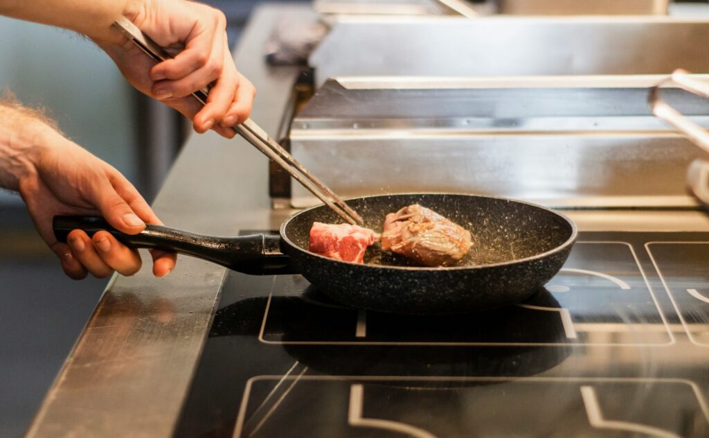 Une personne utilise des pinces métalliques pour faire cuire des morceaux de viande dans une poêle à frire noire sur une cuisinière, un peu comme une aide ménagère du CESU préparant des repas dans une cuisine bien équipée.