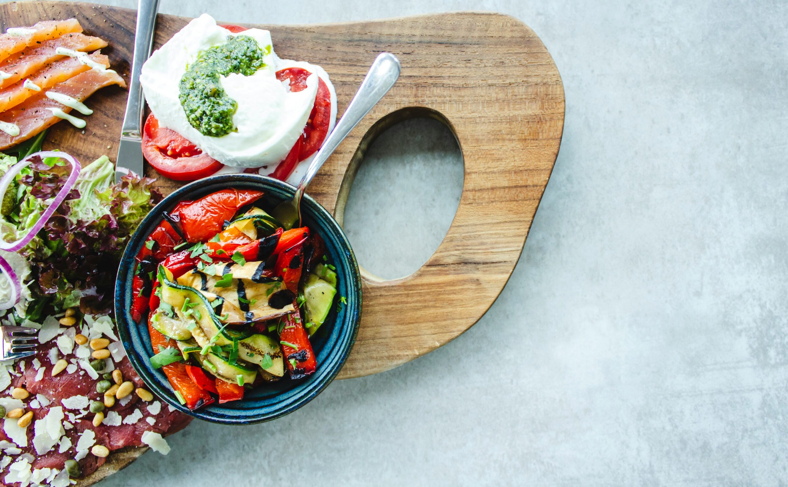 Une planche en bois présente un bol de légumes grillés, une salade fraîche avec des légumes verts et des noix, et des tranches de tomates garnies de mozzarella et de pesto. Des tranches de saumon se trouvent à proximité. L'arrière-plan est une surface de pierre grise.
