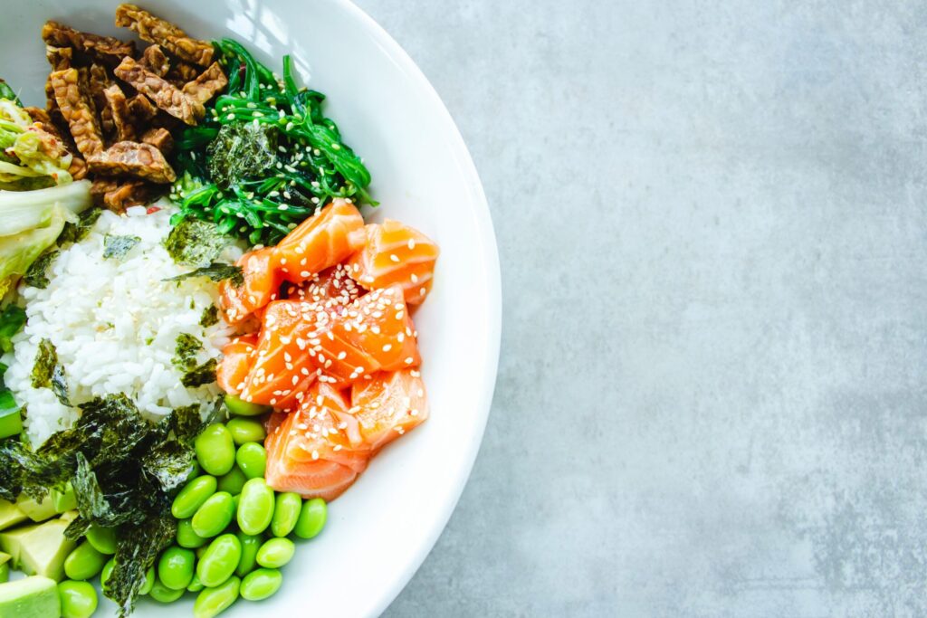 Un bol blanc rempli de riz, de tranches de saumon cru garnies de graines de sésame, d'edamame, d'algues, d'avocat, de légumes verts et de tempeh est posé sur une surface gris clair - parfait pour un repas sain après avoir organisé les services d'aide ménagère du CESU.
