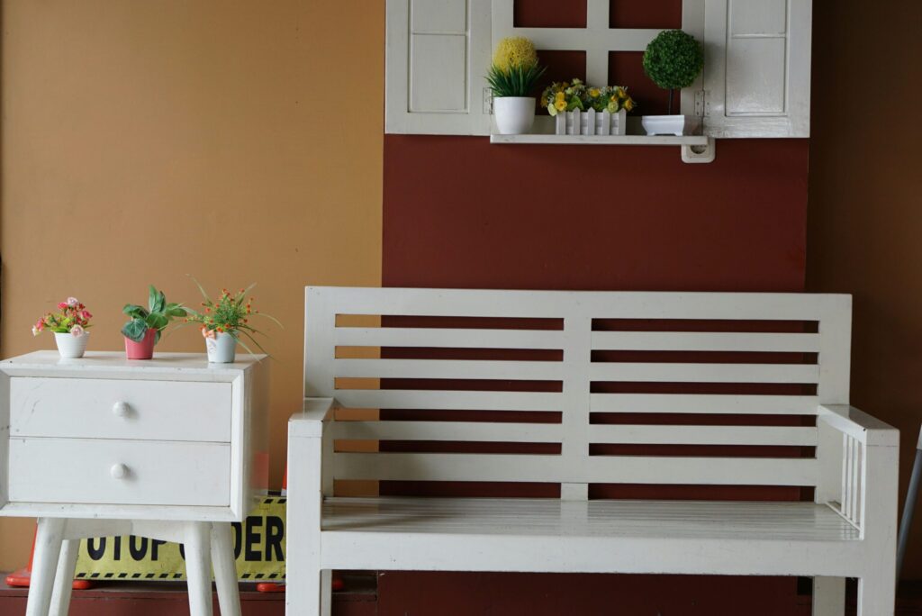 Un banc en bois blanc se trouve à côté d'une petite table blanche avec des plantes en pot. Au-dessus, une fausse fenêtre avec d'autres plantes décore un mur bicolore brun et rouge. Un panneau "Attention" est partiellement visible derrière la table.