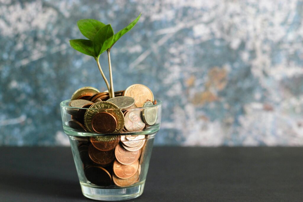 Une petite plante verte poussant dans un verre transparent rempli de pièces de monnaie assorties, sur un fond flou bleu et gris.