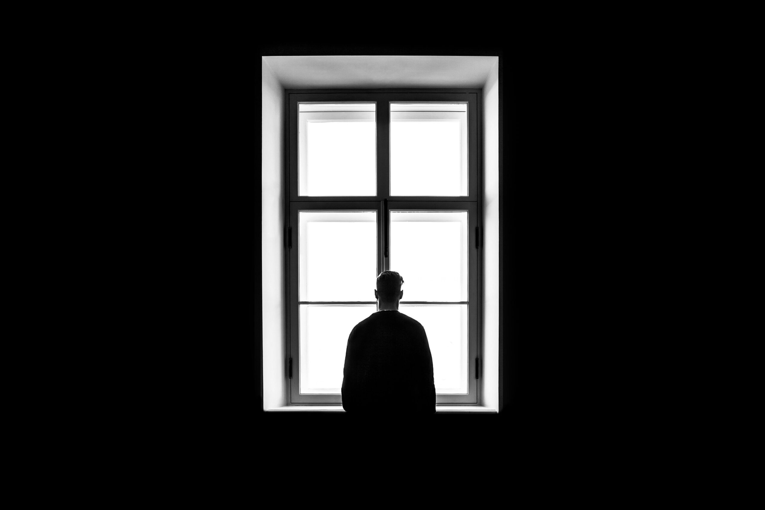 Une personne est assise, seule, en silhouette, face à une grande fenêtre éclairée par une lumière vive à l'extérieur, entourée d'obscurité.