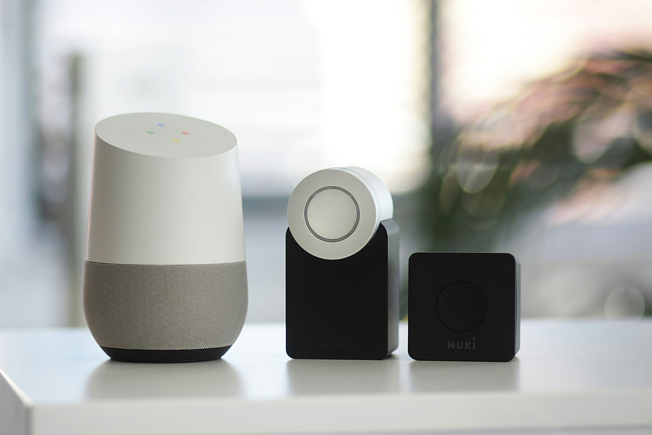 Une enceinte intelligente Google Home est posée sur une surface blanche à côté de deux serrures intelligentes Nuki noires, le tout sur un fond flou.