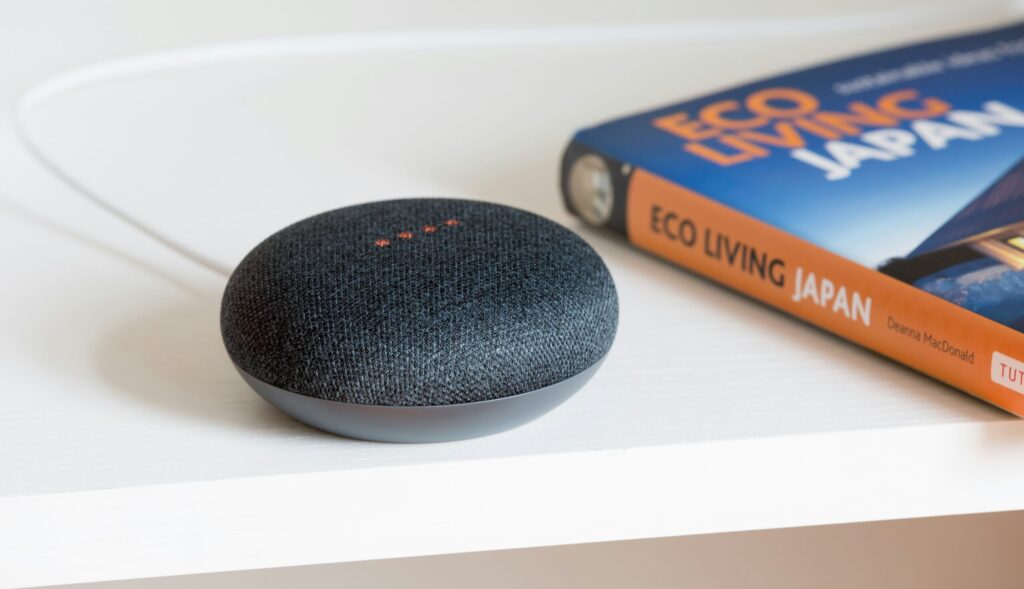 Un haut-parleur intelligent avec un dessus en tissu foncé est posé sur une surface blanche près d'un livre intitulé Eco Living Japan. Un câble blanc est branché sur l'enceinte.