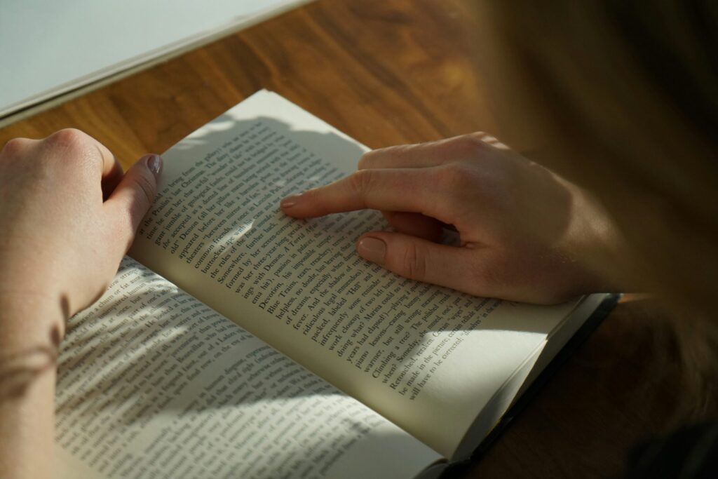 Une personne lit un livre sur une table en bois, en utilisant son index droit pour suivre le texte sur la page. La lumière douce du soleil projette des ombres sur le livre et les mains.