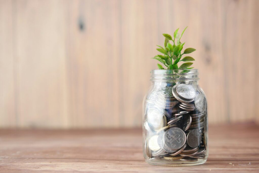 Une petite plante verte pousse au sommet d'un bocal en verre rempli de pièces de monnaie, placé sur une surface en bois sur un fond en bois, symbolisant la croissance financière et l'épargne.