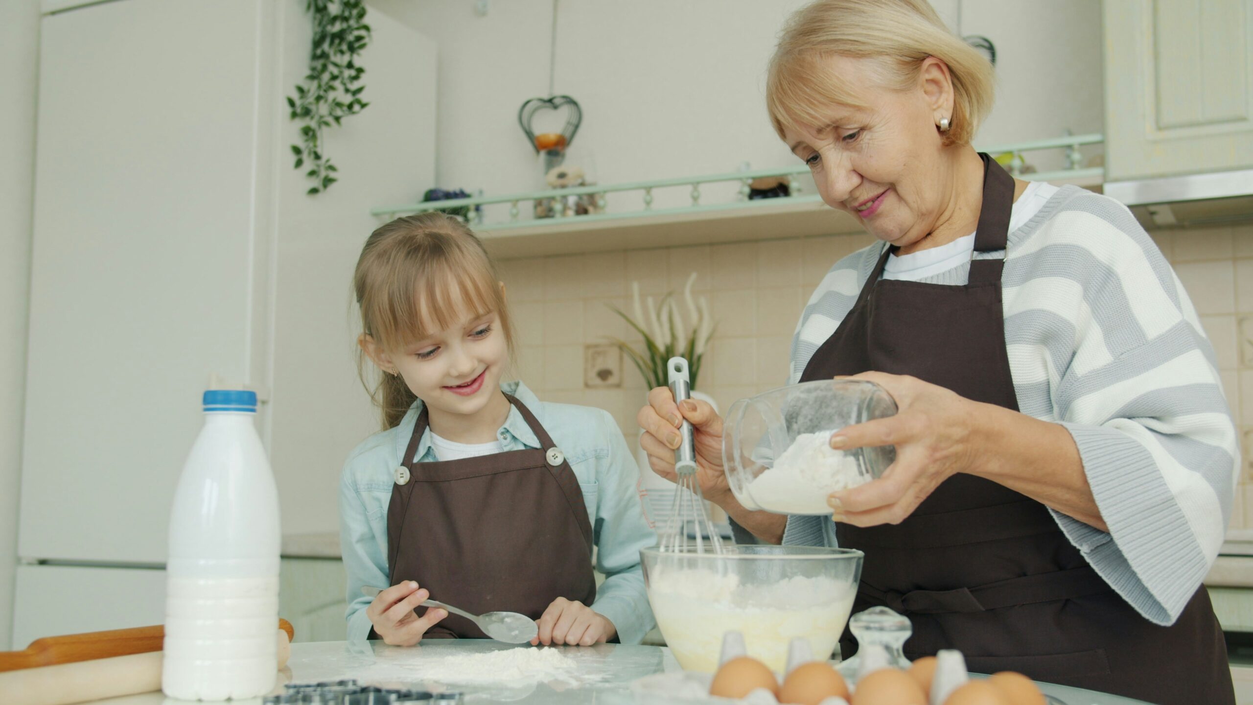 Une femme âgée souriante et une jeune fille, toutes deux vêtues d'un tablier, se tiennent dans une cuisine et mélangent des ingrédients dans un bol. Des œufs, du lait et des ustensiles de cuisine sont posés sur le comptoir pendant qu'elles cuisinent ensemble.