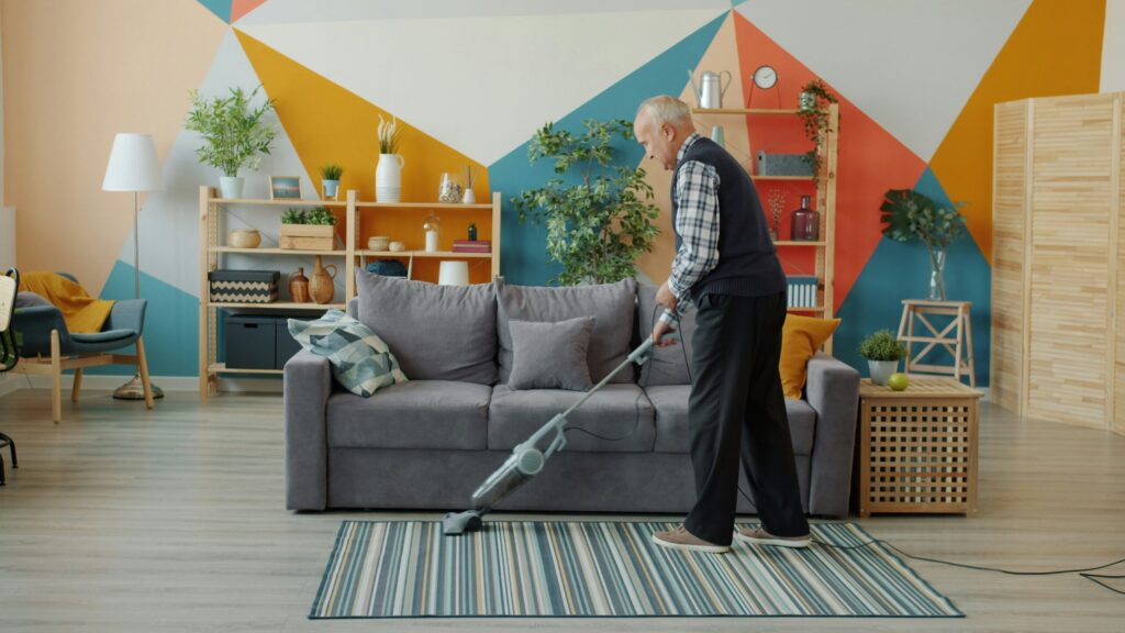 Un homme âgé passe l'aspirateur sur un tapis rayé dans un salon moderne avec un canapé gris, des plantes, des étagères et un mur géométrique coloré en arrière-plan.