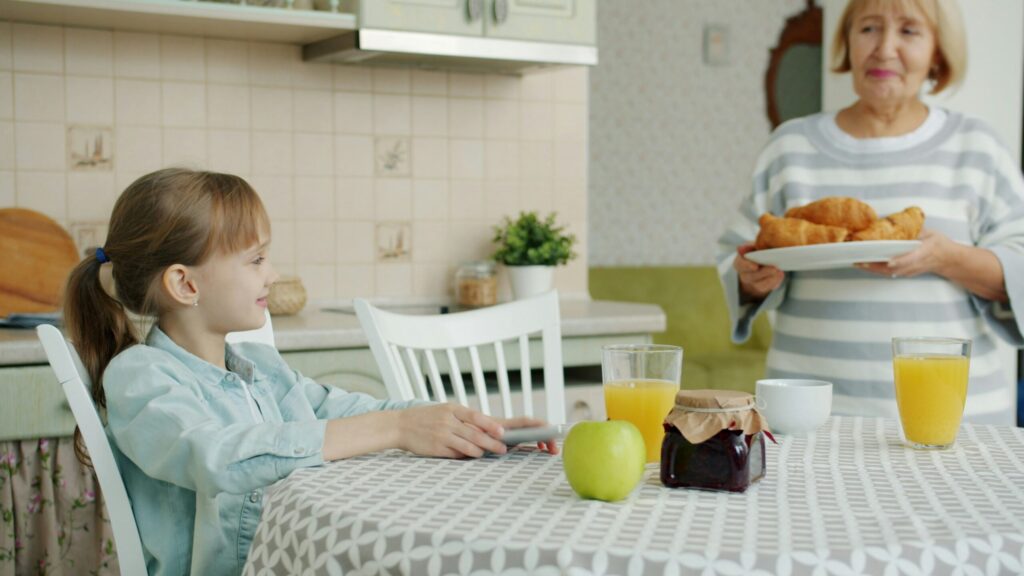 Une jeune fille est assise à une table de cuisine avec un jus de fruit, une pomme et de la confiture. Une femme plus âgée se tient à proximité, tenant une assiette de croissants et souriant à la jeune fille. L'arrière-plan de la cuisine est lumineux et confortable.