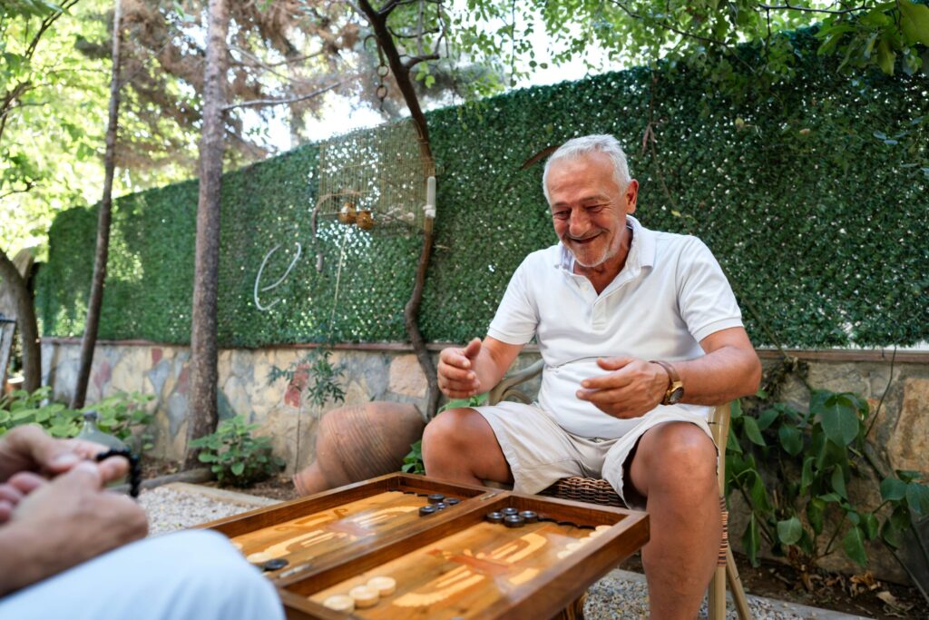 Un homme âgé sourit et fait des gestes tout en jouant à un jeu de société en plein air avec son aide ménagère CESU, assise à une table en bois. La verdure luxuriante et un mur de pierre entourent le jardin accueillant.