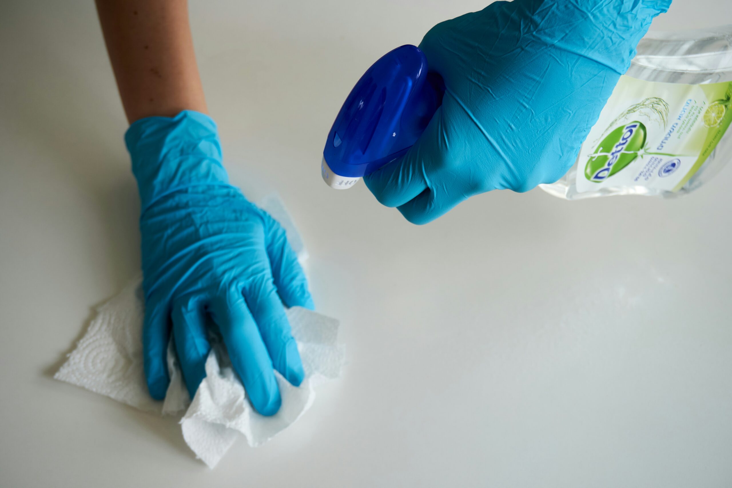 Une personne portant des gants bleus vaporise du nettoyant à partir d'une bouteille et essuie une surface blanche avec une serviette en papier, assurant ainsi une protection contre les punaises de lit.
