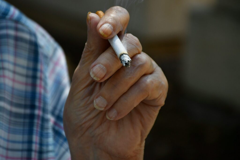 Gros plan sur la main d'une personne, les ongles jaunis saisissant une cigarette allumée, la fumée étant faiblement visible. La personne porte une chemise à carreaux - des mains qui pourraient plus tard être utilisées pour nettoyer un Thermomix.