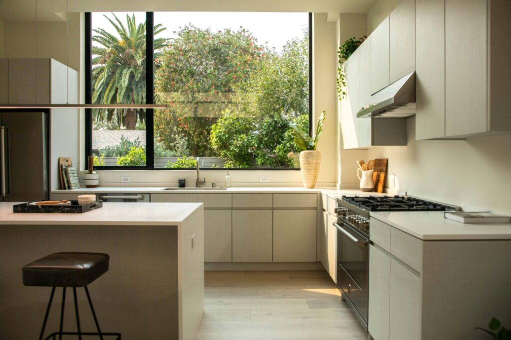 Une cuisine moderne avec des armoires de couleur claire, un îlot central avec un tabouret et une cuisinière à gaz offre un espace accueillant pour préparer des repas sains et prévenir la dénutrition, tandis que de grandes fenêtres encadrent la verdure luxuriante et les palmiers à l'extérieur.