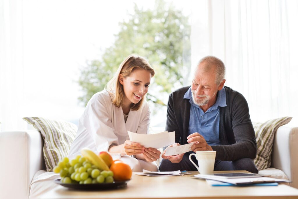 Une jeune femme et un homme plus âgé sont assis ensemble sur un canapé, souriant et regardant des documents sur la façon de nettoyer un Thermomix. Une table devant eux contient des fruits, une tasse et des documents. La pièce est baignée d'une lumière naturelle intense.